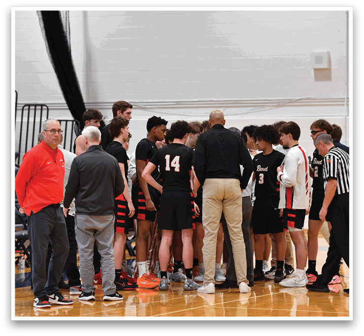 A group of young men are gathered on a basketball court, wearing black and red uniforms. They are standing in a circle, engaged in a conversation with a referee. AI generated content