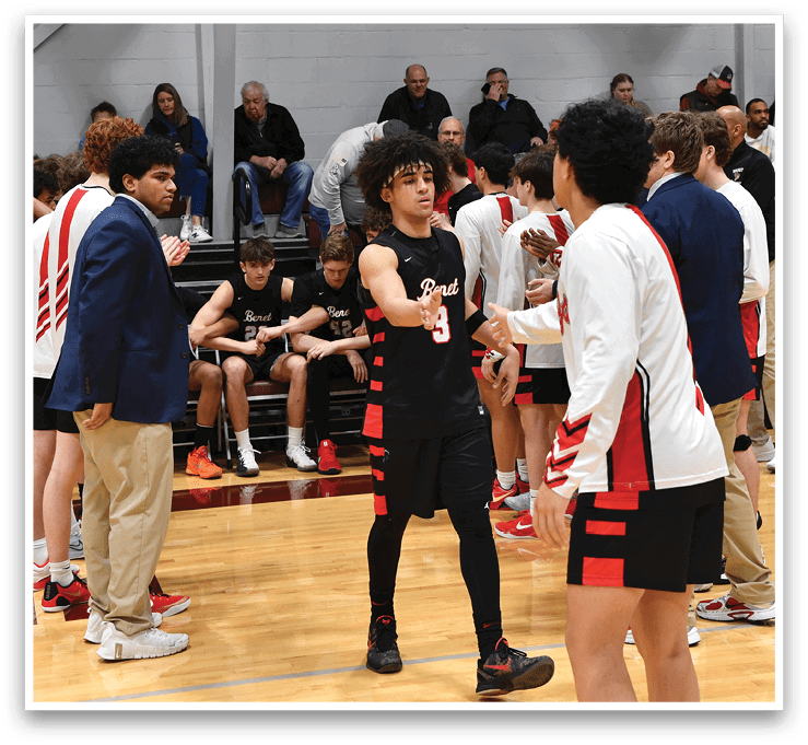 A group of people are standing on a basketball court, some of them wearing red and black uniforms. They are engaged in a conversation, possibly discussing the game or strategizing. There are several chairs placed around the court, and a laptop can be seen on one of them. The people are standing in various positions, some closer to the camera and others further away, creating a dynamic scene. AI generated content