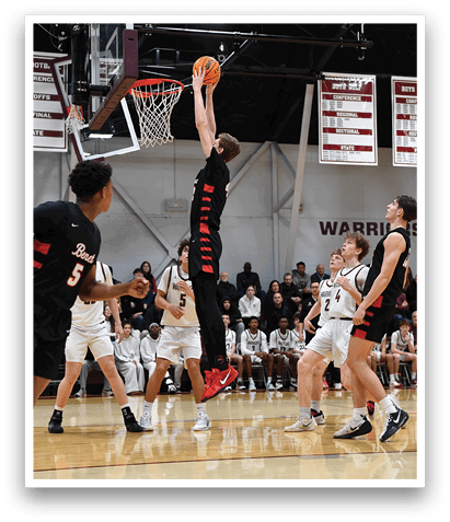 A basketball game is in progress with players on the court. One player is jumping to block a shot, while others are running and playing defense. The crowd watches the game intently. AI generated content