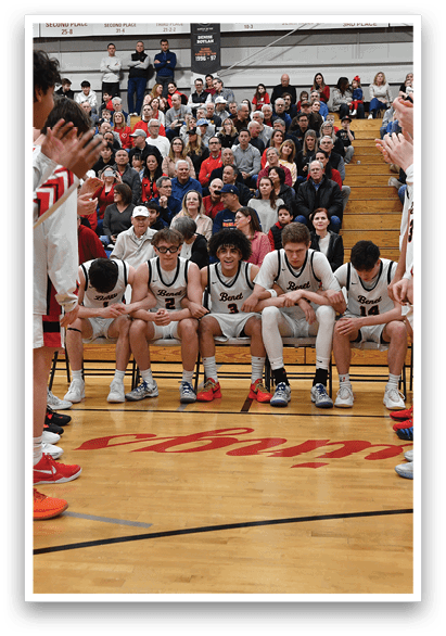 A group of young men are standing on a basketball court, some of them wearing white uniforms. They are holding their hands up in the air, possibly celebrating a victory or preparing for a game. The crowd is watching them, and there are several chairs placed around the court. AI generated content