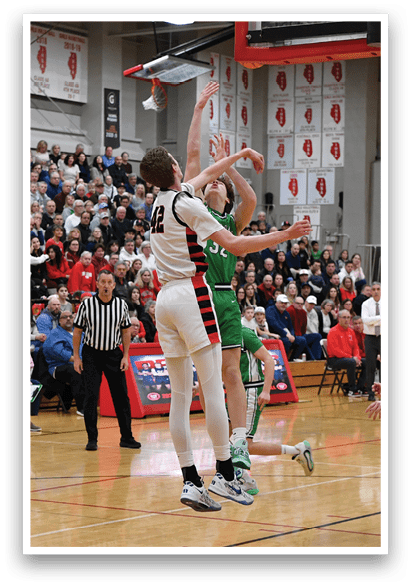 A basketball game is taking place with a crowd watching. The players are wearing uniforms and one player is attempting to block a shot. The crowd is composed of various people, some sitting on chairs and others standing. The atmosphere is lively and competitive. AI generated content