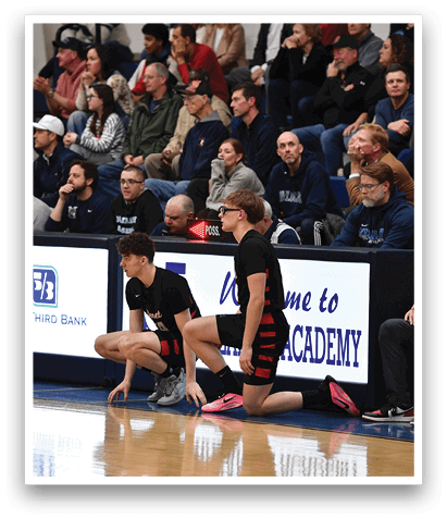 Two men are kneeling on a basketball court, with one of them wearing a red shirt. They are surrounded by a crowd of people, some of whom are sitting on chairs. The scene appears to be a basketball game or practice. AI generated content