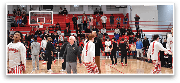 A basketball game is taking place in a crowded gymnasium. The players are wearing red and white uniforms, and there are spectators watching the game. The gym is filled with people, and there are several chairs placed around the area. The atmosphere is lively, and the players are actively engaged in the game. AI generated content