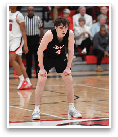 A basketball player is standing on the court, wearing a black shirt and white shoes. He is looking down at the ground, possibly preparing to make a move. There are several other players on the court, some of them wearing red and white uniforms. A referee is also present, watching the game closely. The scene takes place in a gymnasium, with a crowd of spectators watching the game. AI generated content