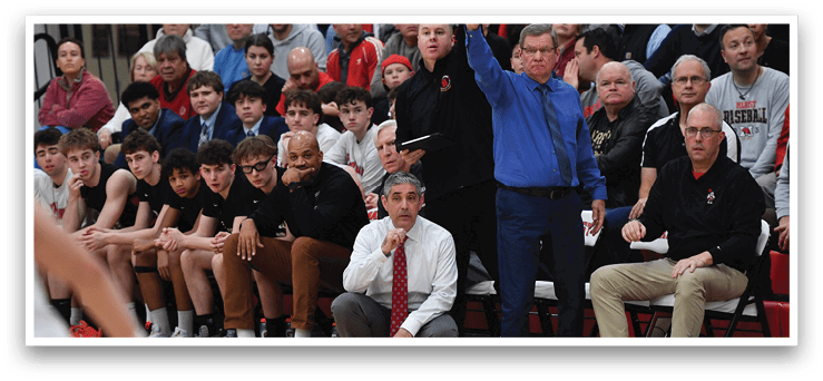 A basketball game is taking place with a man in a blue shirt and khaki pants sitting on a chair, watching the game. The man is wearing a tie and appears to be the coach. There are several other people in the scene, some of whom are also sitting on chairs, while others are standing. A basketball player is holding his arms up in the air, possibly celebrating a point. AI generated content
