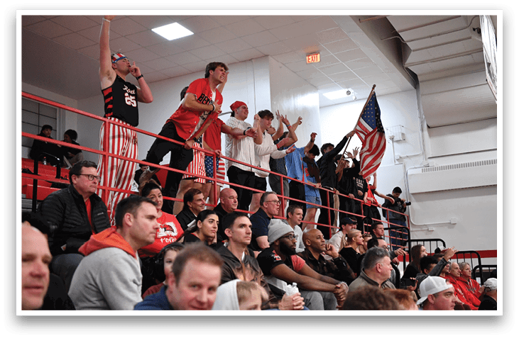 A group of people are sitting in the stands, watching a game. They are wearing red, white, and blue clothing, and some are holding American flags. The crowd is engaged and excited, cheering for their team. AI generated content