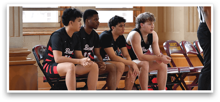 A group of boys are sitting on chairs in a gym, waiting for their turn to play basketball. A referee is standing nearby, watching over the game. AI generated content