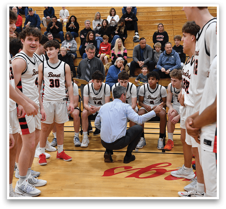 A group of young men in uniforms are standing on a basketball court, with one man kneeling down to talk to another. The group is composed of both players and coaches, and they are all wearing uniforms. The players are standing in various positions around the court, while the coaches are gathered in the middle. The scene appears to be a casual conversation between the players and the coaches. AI generated content