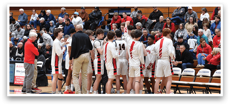 A group of young men are on a basketball court, some of them wearing red and white uniforms. They are standing in a circle, possibly discussing their game strategy or listening to a coach. The crowd of spectators is watching the game from the stands, and there are several chairs placed around the court. AI generated content