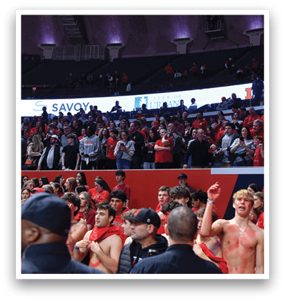 A group of people are standing in a stadium, some of them are wearing red shirts. They are watching a game and cheering for their team. AI generated content