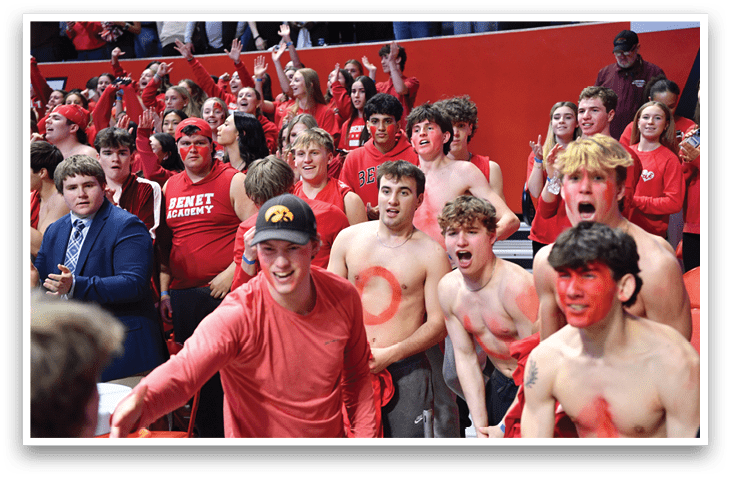 A group of people are sitting in the stands, watching a game. They are wearing red shirts and are painted with red paint. Some of the people are also wearing red hats. The crowd is cheering and clapping, creating a lively atmosphere. AI generated content