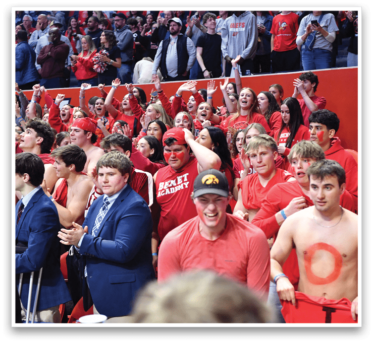 A group of people are sitting in the stands, wearing red shirts and holding up signs. They are cheering for a team, possibly the Fighting Illini. The crowd is diverse, with some people sitting close to the front and others further back. The atmosphere is lively and energetic, as the fans are excited to support their team. AI generated content