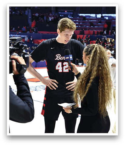 A man and a woman are talking to each other on a basketball court. The woman is wearing a black shirt and has long hair. The man is wearing a basketball jersey and is standing next to the woman. There are several other people in the background, some of whom are holding cameras. AI generated content