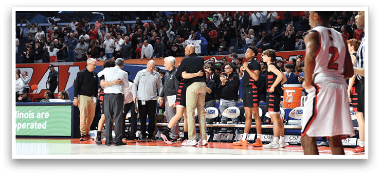 A group of men are standing on a basketball court, with some of them wearing red and white uniforms. They are engaged in a conversation, with one man holding his arm around another. The scene appears to be a game, with a crowd of people watching from the stands. AI generated content