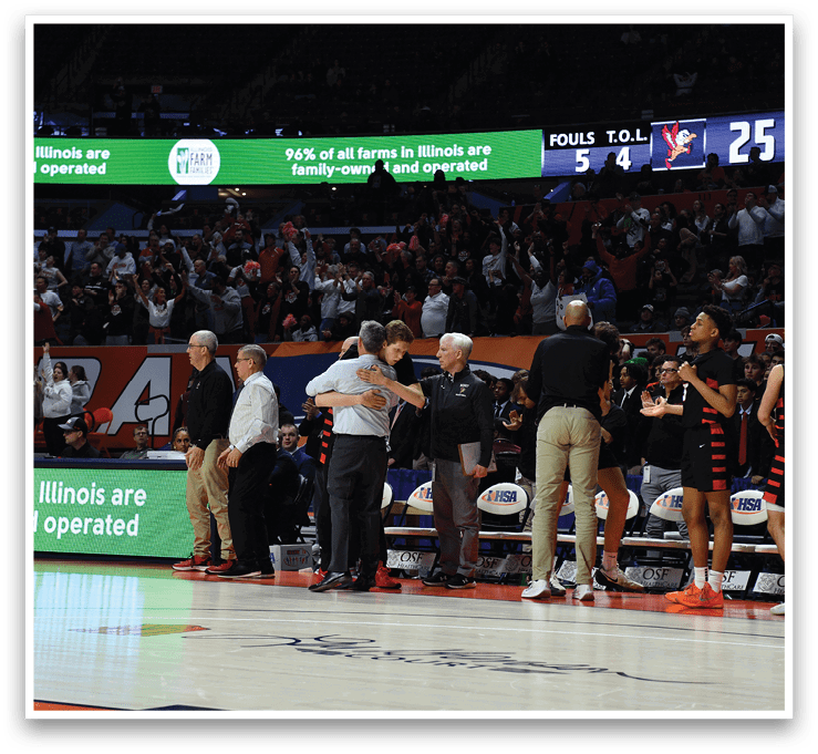 A group of people are standing on a basketball court, with some of them wearing orange and black uniforms. They are holding hands and hugging each other, possibly celebrating a victory or showing support for one another. The crowd of people in the background is watching the event, and there is a clock visible in the upper right corner of the image. AI generated content