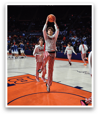 Two basketball players in red and white striped uniforms are playing in a game. One player is jumping to shoot the ball while the other player is running towards the basket. There are several other players on the court, some of them are wearing white uniforms. The scene is set in a basketball court with a crowd watching the game. AI generated content