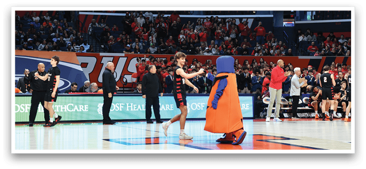 A mascot in an orange costume is on the court, interacting with a woman in a black dress. The mascot is holding a microphone, and the woman is holding a cell phone. The scene takes place in a basketball court, with several people in the background. AI generated content