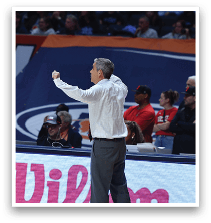 A man is standing on a basketball court, wearing a white shirt and pointing at something. He is surrounded by a crowd of people, some of whom are sitting on chairs. The scene appears to be a game or event taking place in a stadium. AI generated content