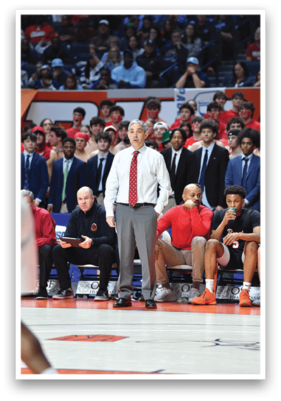 A group of people are sitting on a bench, watching a basketball game. A man in a red tie is standing on the sideline, observing the game. AI generated content
