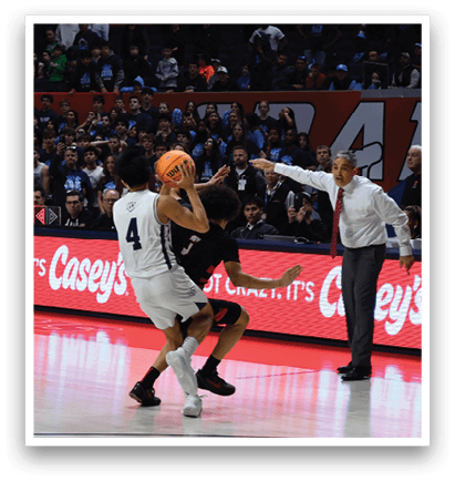 A basketball game is taking place with a crowd of people watching. One player is attempting to shoot the ball while another player tries to block the shot. AI generated content