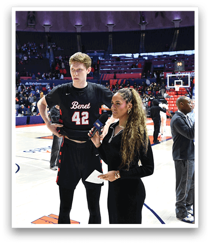 A man and a woman are standing on a basketball court, with the woman holding a microphone. They are surrounded by several other people, some of whom are holding cell phones. The scene appears to be a press conference or interview. AI generated content