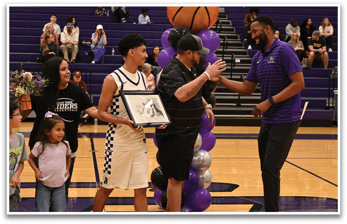A group of people, including a young man holding a framed item, are standing on a basketball court. A man in a black shirt and hat is shaking hands with the young man, while another man in a purple shirt stands nearby. There are several other people in the scene, some of whom are sitting on chairs. A basketball can be seen in the background. Contenido generado con IA