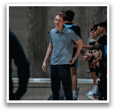A man wearing a gray shirt and black pants stands on a basketball court, smiling. He is surrounded by other people, some of whom are wearing black shirts. AI generated content