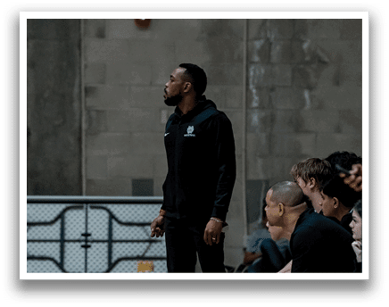 A group of people are sitting on a bench in a gym. A man in a black shirt is standing in front of them. AI generated content