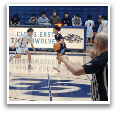 A basketball game is taking place with a group of people on the court. A referee is present, and a player is dribbling the ball. There are several other players on the court, some of whom are sitting on chairs. The scene is set in a gym with a blue floor. AI generated content
