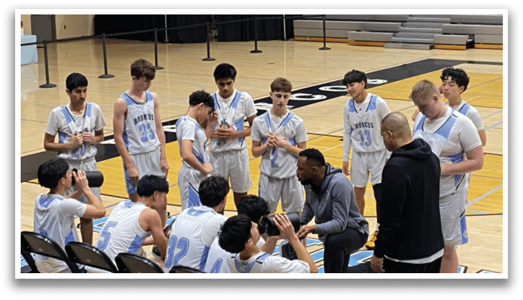 A group of young men in uniform are gathered on a basketball court, some sitting and others standing. They are wearing blue and white uniforms and are likely discussing their game strategy. AI generated content