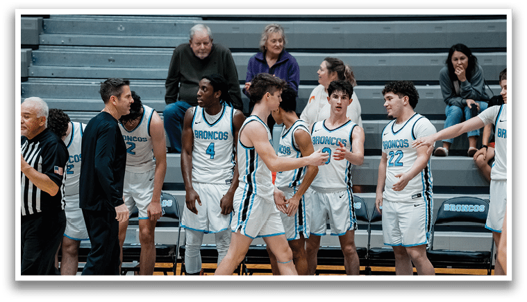 A group of young men are playing basketball, with one team standing on the court and the other team sitting on a bench. The players on the court are wearing blue and white uniforms, while the players on the bench are wearing orange and white uniforms. The players on the court are holding their hands up, possibly celebrating a point or a victory. AI generated content
