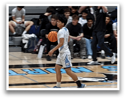 A basketball game is taking place with players on the court and spectators in the stands. A man in a white shirt is holding a basketball while another player in a black shirt is running towards him. The players are actively engaged in the game, and the audience is watching the action unfold. AI generated content