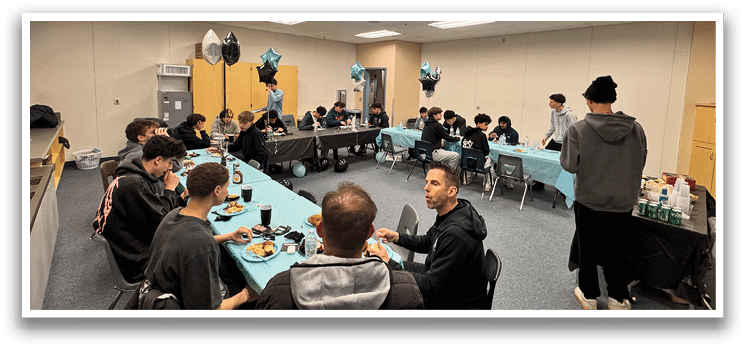 A group of people are sitting around a table in a room with a blue tablecloth. The table is set with plates, cups, and utensils. There are balloons on the table, and a cake is also present. The people are engaged in conversation and enjoying their time together. AI generated content