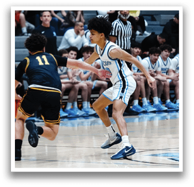 A basketball game is in progress with players on the court. A boy is running with the ball while another player tries to block him. The players are wearing uniforms and the game is being watched by a crowd of people. AI generated content