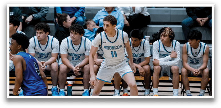 A group of young men are playing basketball, with one man in a white uniform standing in front of the others. The players are wearing uniforms with the word “Broncos" on them. The players are sitting on a bench, and there are water bottles and a backpack on the bench as well. AI generated content