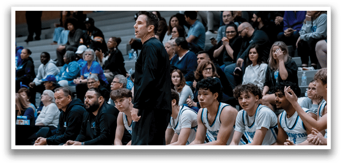 A man is standing on a basketball court, talking to a group of young men. They are all wearing uniforms, and some are sitting on chairs. The man appears to be a coach, giving instructions to the players. AI generated content