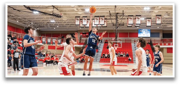 A basketball game is in progress with a player jumping to make a basket. The player is wearing a blue shirt and is in the air with the basketball in his hand. The game is being played in a gymnasium with a crowd of spectators watching the action. AI generated content