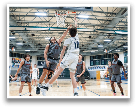 A basketball game is in progress with players on the court. One player is jumping to block a shot, while another player is trying to score. The game is being played indoors, and there are several other players on the court. AI generated content