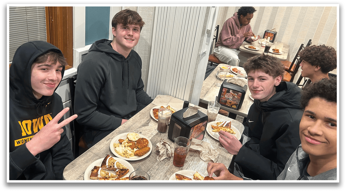 Four young men are sitting around a dining table, enjoying a meal together. They are smiling and posing for a photo, with a pizza in front of them. There are several cups and a fork on the table, indicating that they are eating and drinking. The atmosphere appears to be friendly and relaxed, as they share a pleasant dining experience. AI generated content