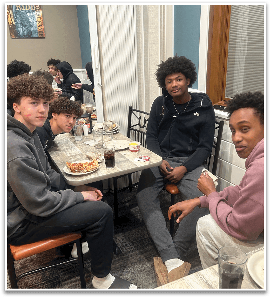 Four young men are sitting around a dining table, enjoying a meal together. They are surrounded by various items such as cups, forks, knives, and a pizza. There are also chairs placed around the table, and a clock on the wall. The atmosphere appears to be casual and friendly, as the group shares a meal and spends time together. AI generated content