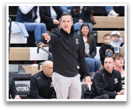 A man in a black and gold jacket is standing on a basketball court, pointing at something. He is wearing a black hat and a black and gold jacket. There are several other people in the scene, including a man sitting in a chair and another man standing. A sports ball is visible on the court. AI generated content