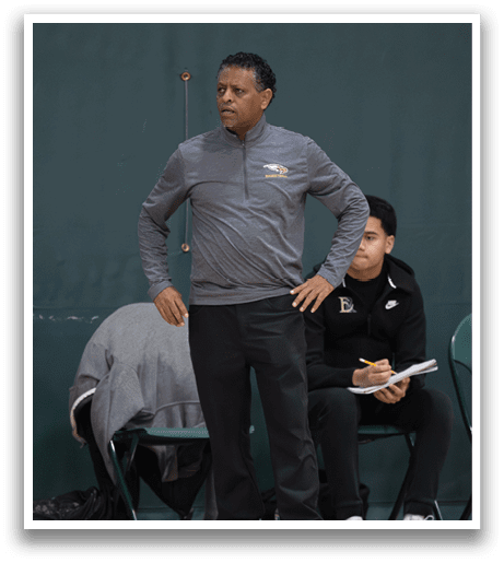 A man wearing a gray jacket and black pants stands on a basketball court, looking at a player sitting on a chair. AI generated content