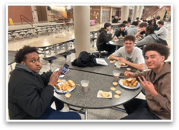 Three young men are sitting at a table in a cafeteria, eating food and drinking milk. They are using their cell phones while enjoying their meal. AI generated content