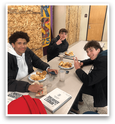 Three young men are sitting at a table, eating food and drinking from cups. They appear to be enjoying their meal together. AI generated content
