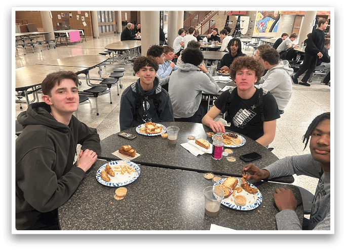 Four young men are sitting around a dining table, enjoying a meal together. They are surrounded by various food items such as sandwiches, cups, and a cell phone. The table is set with plates, forks, knives, and spoons, indicating a proper meal setting. The atmosphere appears to be casual and friendly, as the young men share a meal together. AI generated content