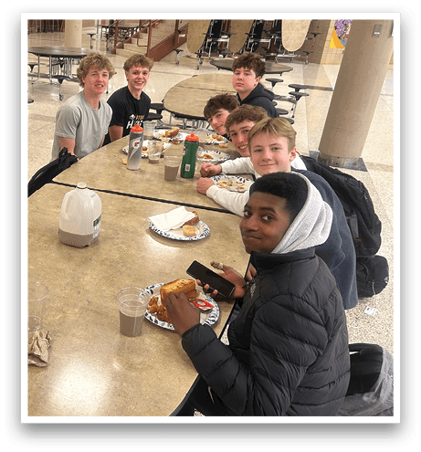 A group of young people are sitting around a table in a cafeteria, eating and drinking. AI generated content