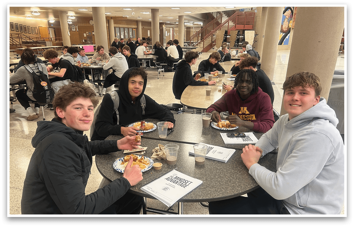 Four young men are sitting around a table, eating food and drinking from cups. They are in a cafeteria setting, surrounded by other people. AI generated content