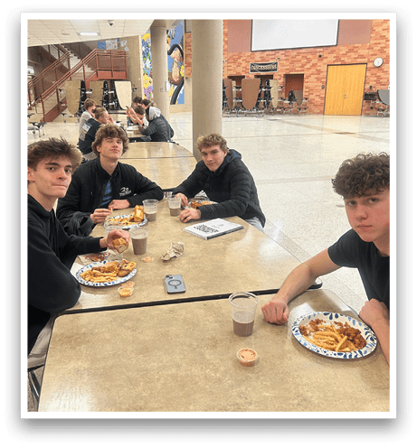 Four young men are sitting around a dining table, enjoying a meal together. They are surrounded by various items such as cups, forks, knives, and spoons. There are also books and a backpack on the table. The scene appears to be a casual gathering of friends sharing a meal. AI generated content