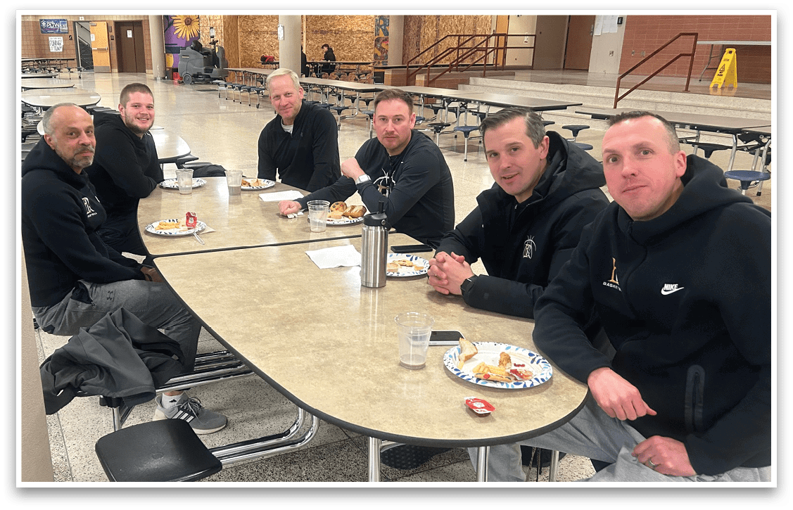 A group of men are sitting around a table in a cafeteria, eating and drinking. AI generated content