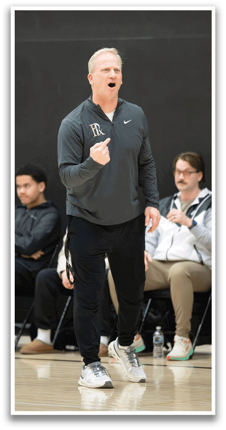 A man wearing a gray sweatshirt stands on a basketball court, holding his hands up in the air. Another man sits in a chair nearby, watching the game. AI generated content
